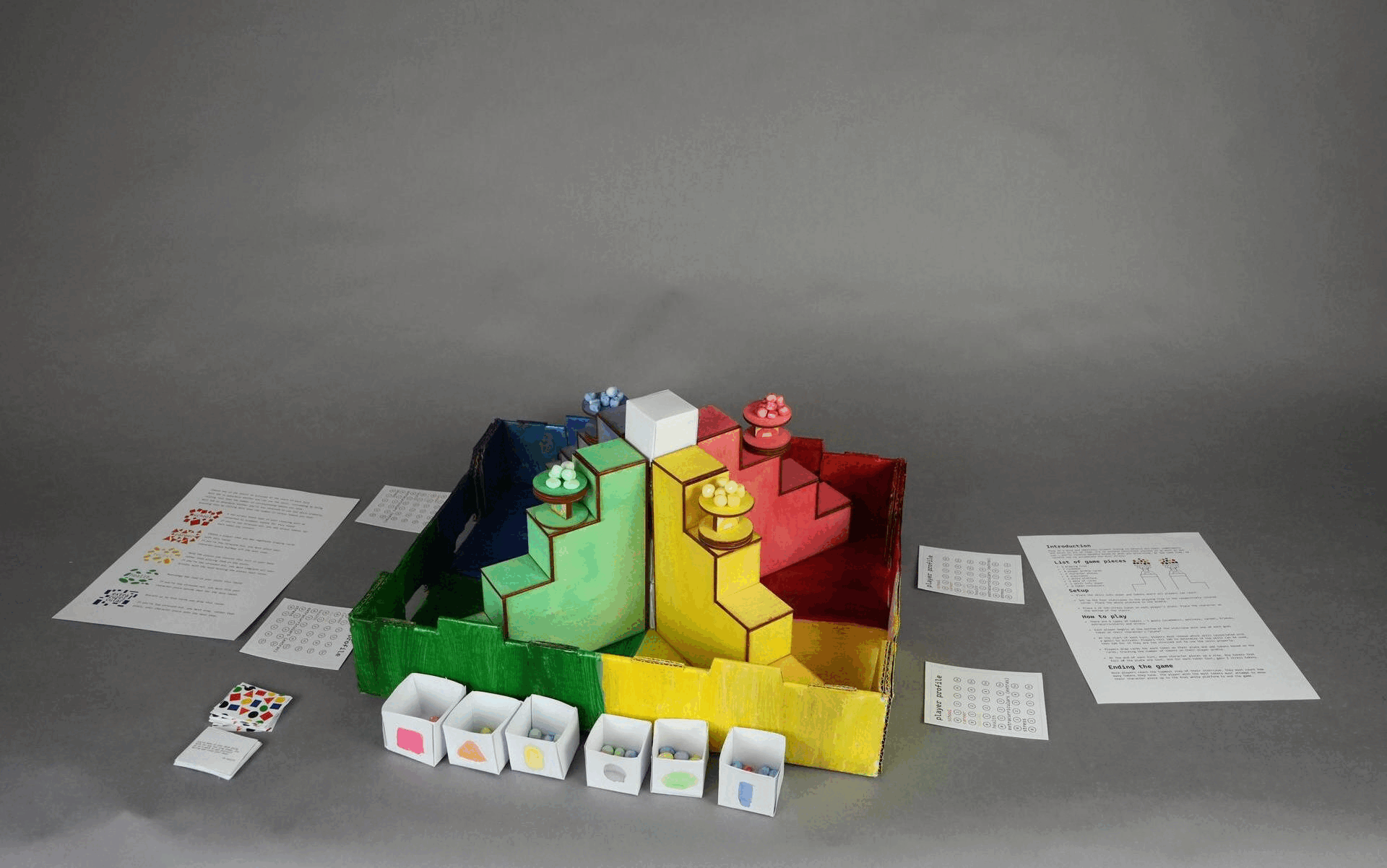 This gif swaps between several photos of a board game. This board game is made up of 4 staircases, colored red, blue, yellow, and green, that converge to a white platform. The character pieces placed on the staircase are students carrying plates, and there are clay tokens of different shapes on their plates.